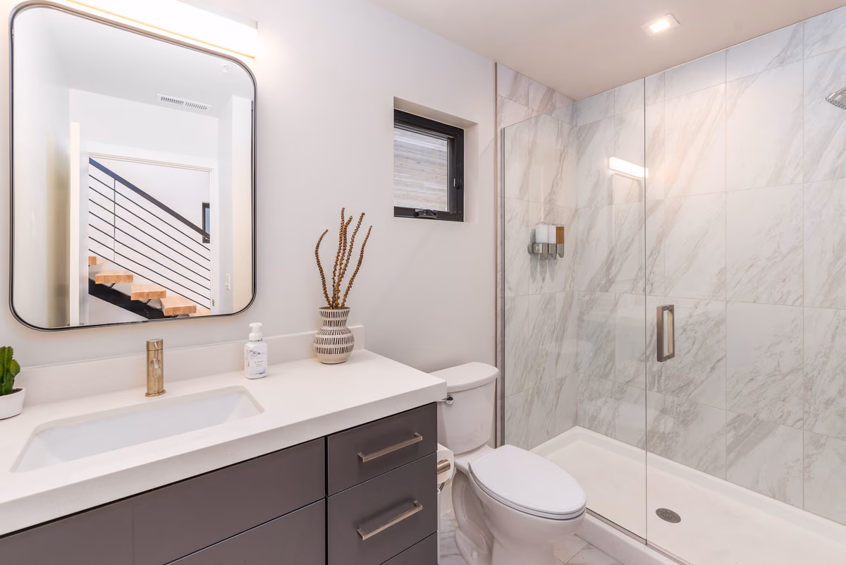 Bathroom with a floating dark vanity and white top, a rounded-corner mirror reflecting the stair, a marble-look tiled shower with a glass door, and a small window.