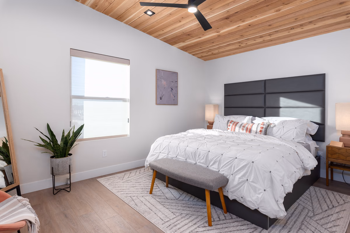 Bedroom with a cedar-plank sloped ceiling and black fan, a dark grey paneled headboard, white bedding, a grey bench, a window, and a patterned rug.