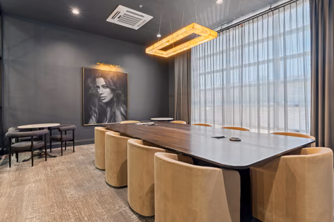 Dark grey board room with a long walnut table, tan upholstered chairs, a framed black-and-white portrait, a glowing rectangular pendant, and sheer curtains over a tall window.