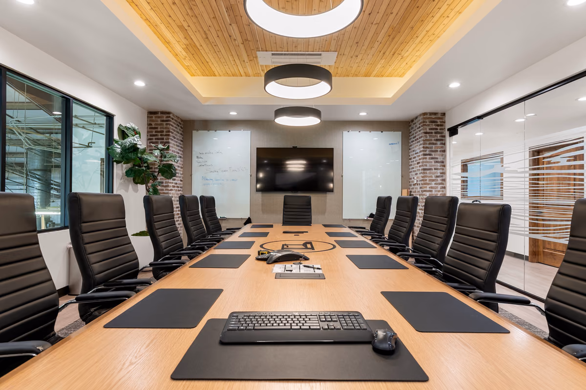 Boardroom seen down the length of an oak table with black leather chairs and desk pads, under a recessed cedar ceiling with round pendants, brick columns, and two glass whiteboards around a wall display.