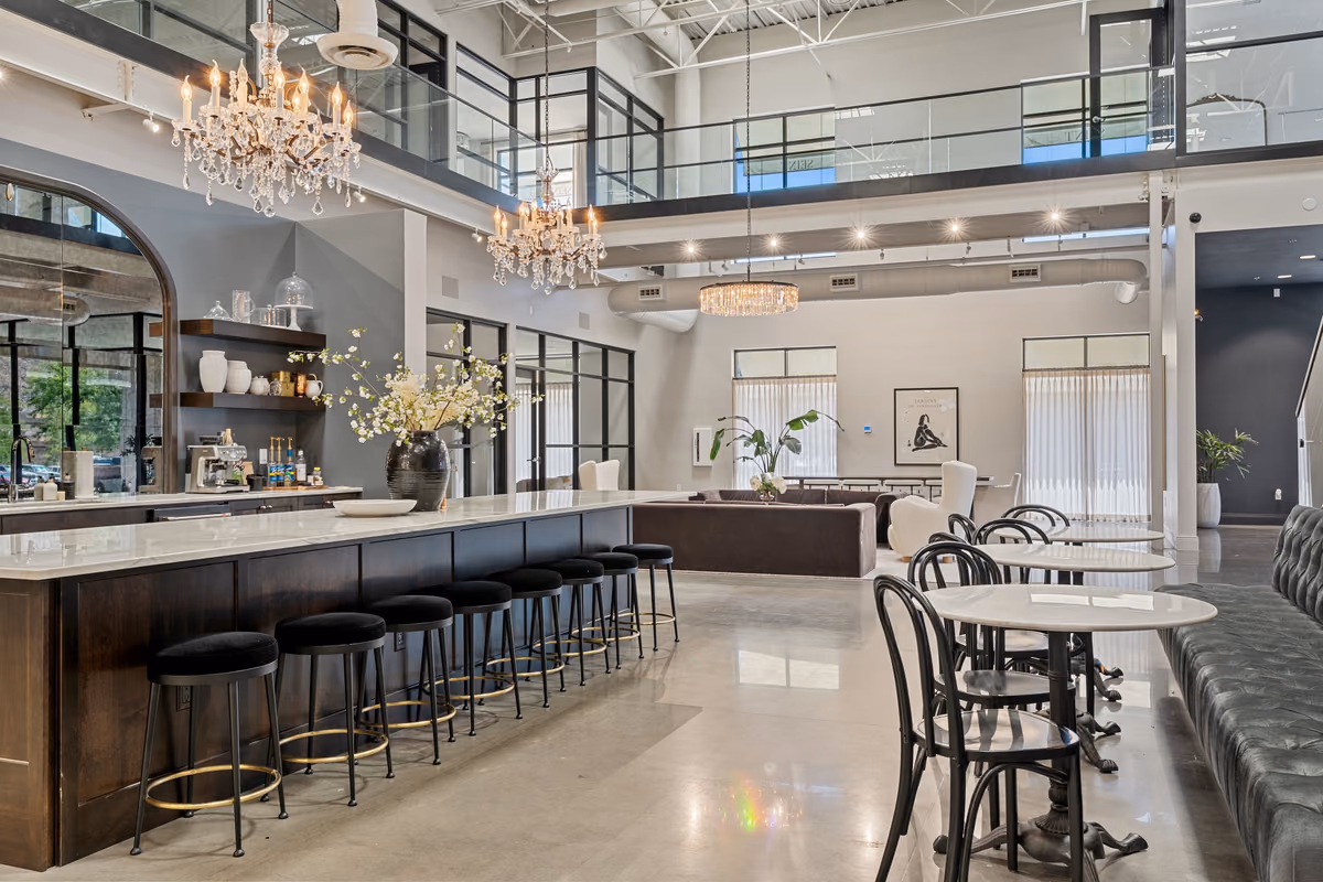 Café island with a white stone top and dark wood base, a row of black stools with brass footrests, crystal chandeliers, a tufted banquette, and a lounge area beyond.