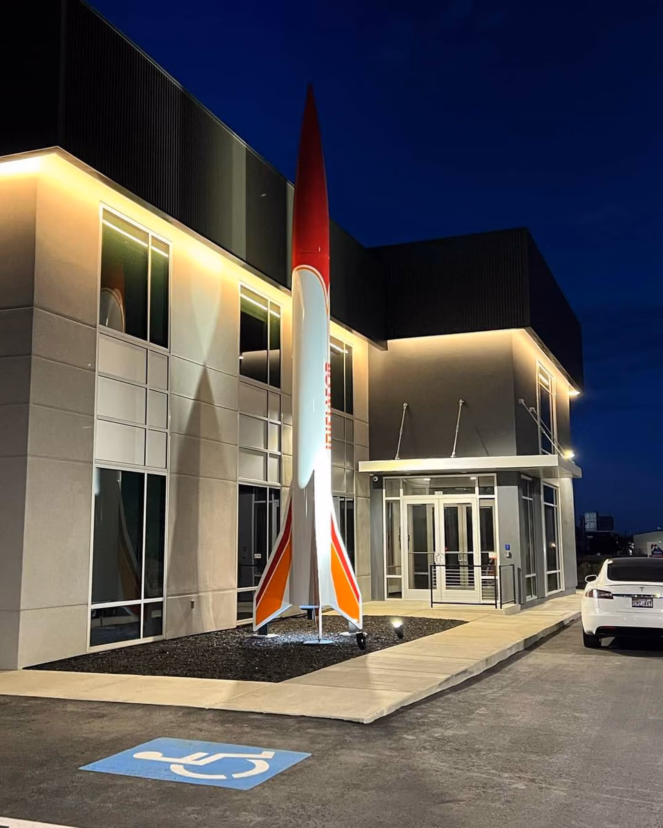 Two-story office building at night with dark metal panel cladding and lit glass entry doors, and a tall white, red, and orange model rocket replica standing in front beside a handicap parking space.