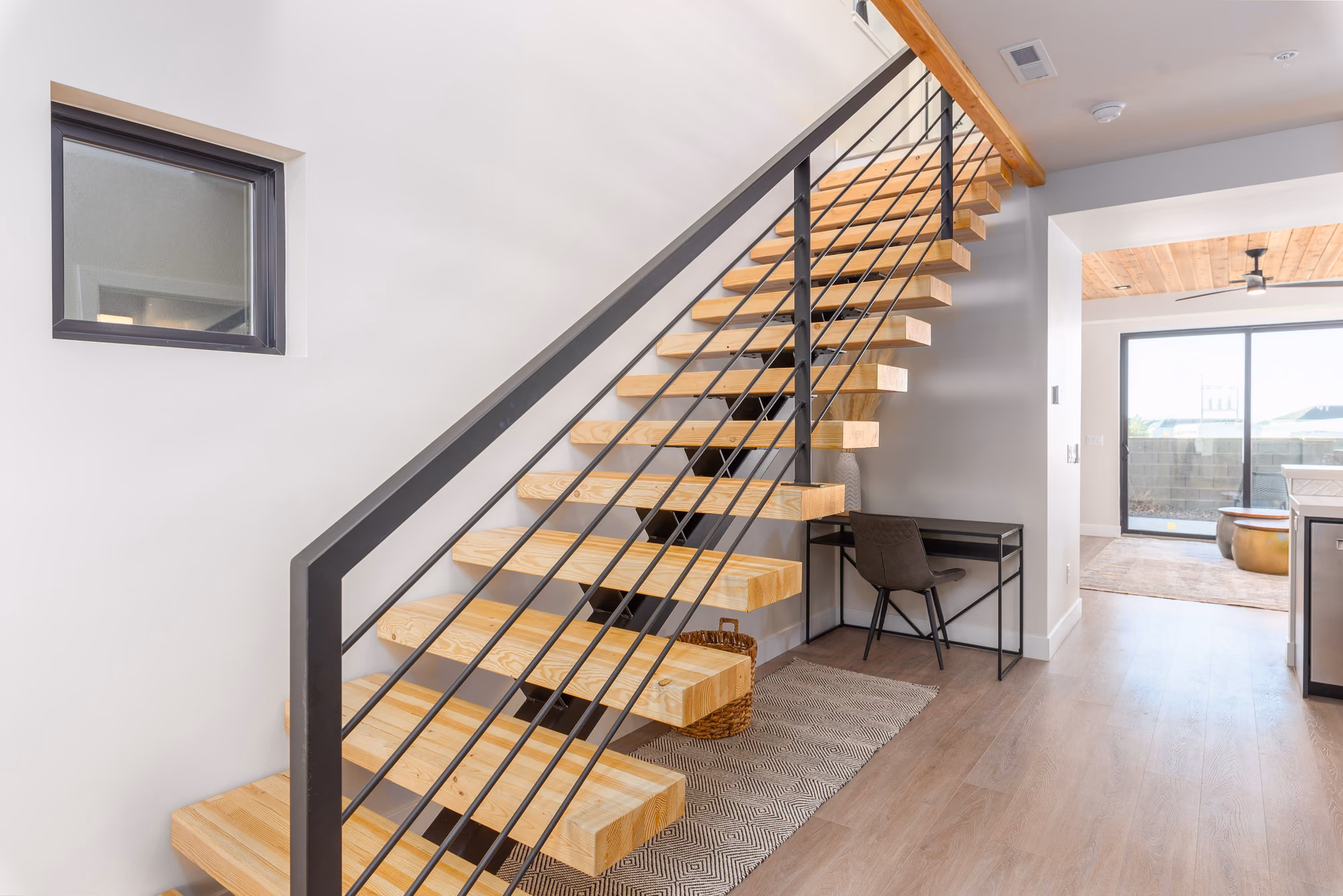 Straight stair with thick timber treads on a black steel stringer and a horizontal rod railing, with a white wall and a desk at the foot of the stair.