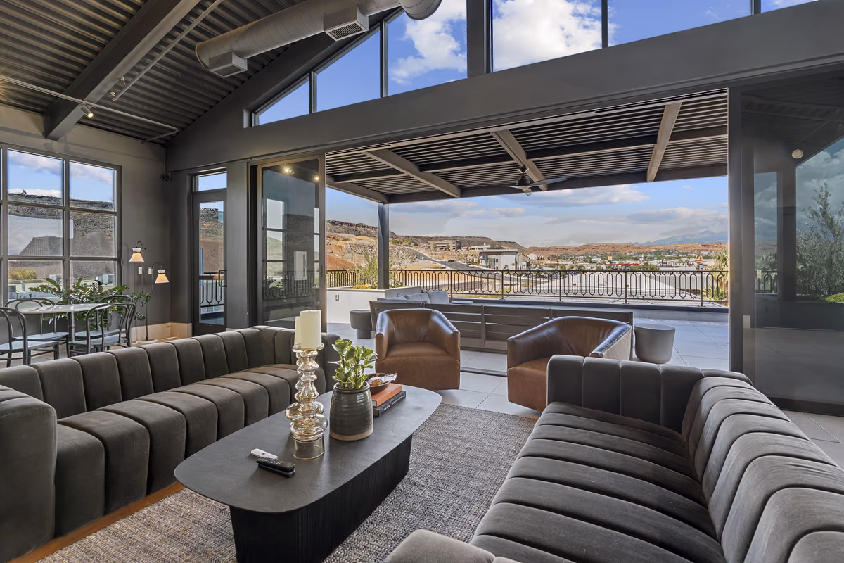 Rooftop lounge seating with grey channel-tufted sofas and leather chairs, looking through open glass sliding doors to a covered deck, an iron railing, and the city and red rock cliffs beyond.