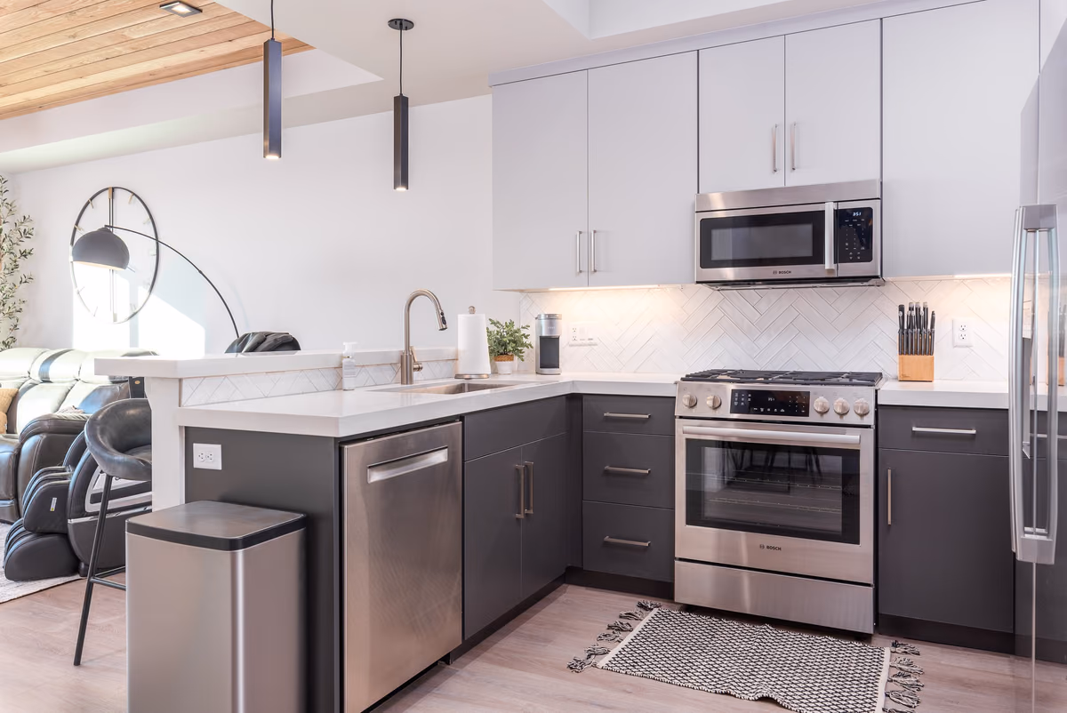 Kitchen with light flat-panel upper cabinets, dark lower cabinets, a white herringbone tile backsplash, stainless appliances, a white counter with a bar, and pendant lights.