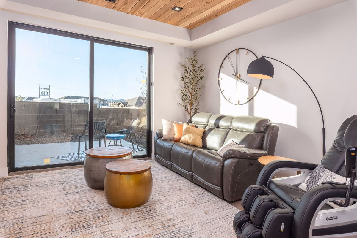 Living room with a cedar-plank ceiling, a sliding glass door to a patio, a dark leather sofa, two round hammered-metal ottomans, a large wall clock, and an arc floor lamp.