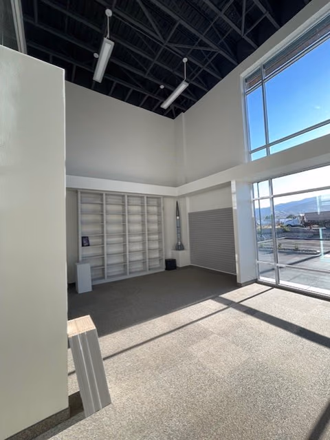 Corner of the two-story room with exposed black roof trusses, tall glass windows looking out toward mountains, and empty white built-in shelving.