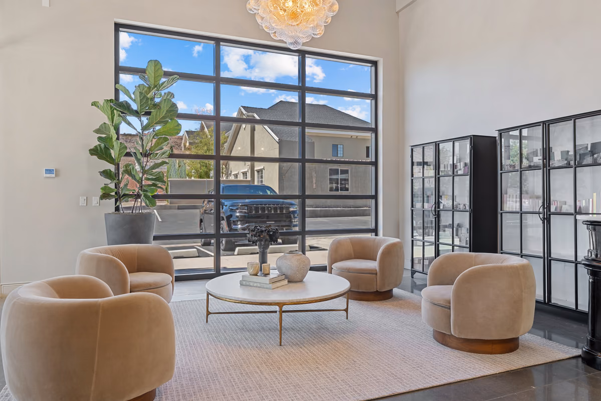 Entry lounge with cream swivel chairs around a round marble table, a large black-framed window onto the parking area, a fiddle-leaf fig, and black glass cabinets.