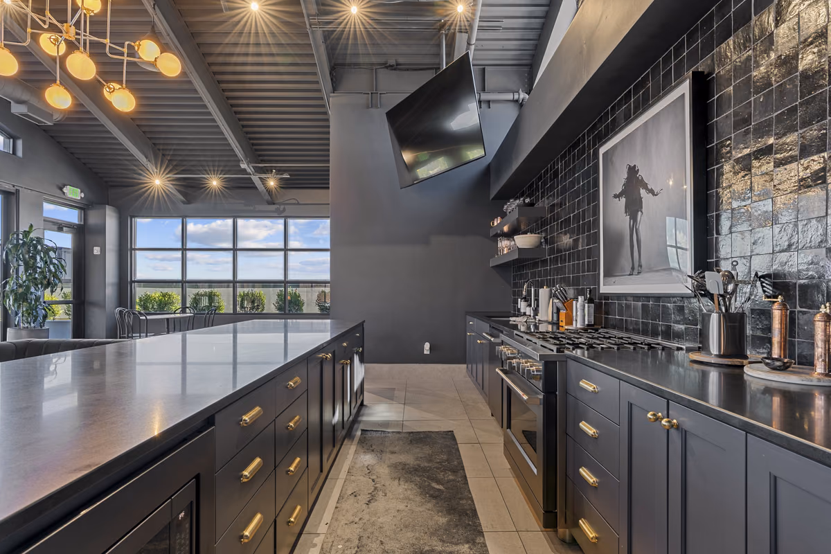 Rooftop kitchen with navy cabinetry and brass pulls, a stainless range, black glazed tile, a dark counter island, a framed black-and-white photograph, and globe pendants under a corrugated ceiling.