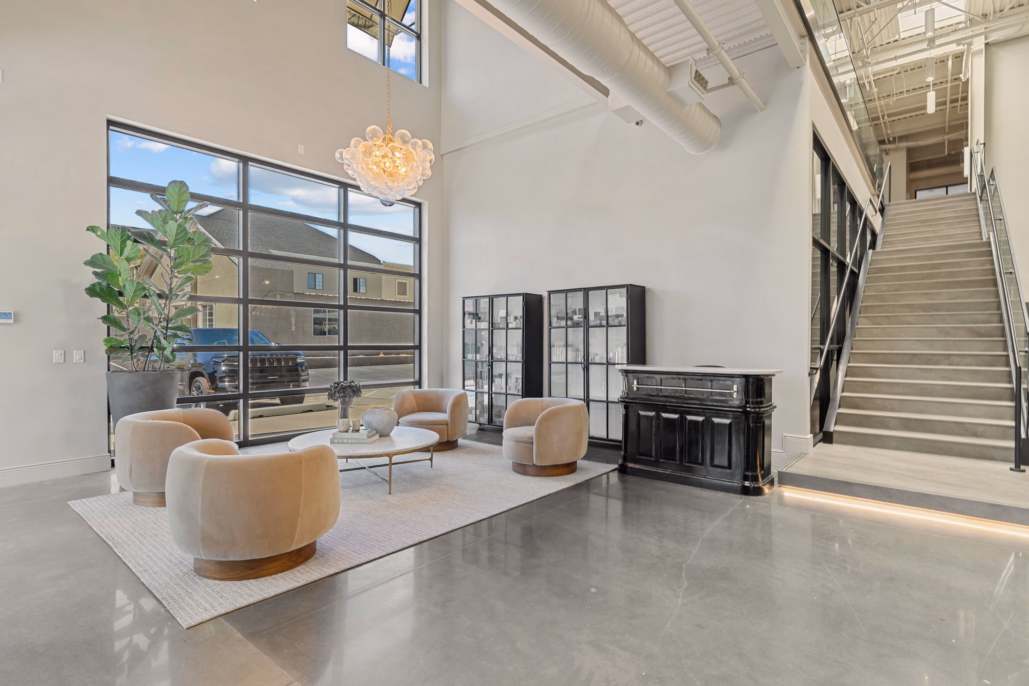 Double-height entry lounge with polished concrete floors, four cream swivel chairs around a round marble table, black glass-fronted cabinets, a bubble chandelier, and a concrete stair with glass and steel rails.