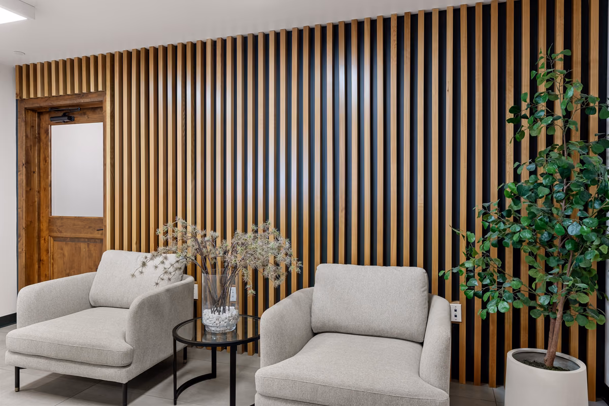 Waiting area with two light grey armchairs, a round glass side table, and a potted tree in front of a wall of vertical wood slats over a dark backing.