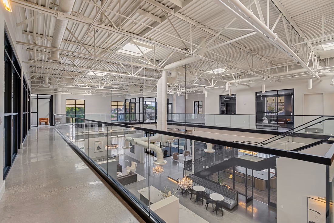 Mezzanine corridor with a polished concrete floor beside a glass guardrail, looking down on the café and banquette, under white steel trusses, skylights, and round ductwork.