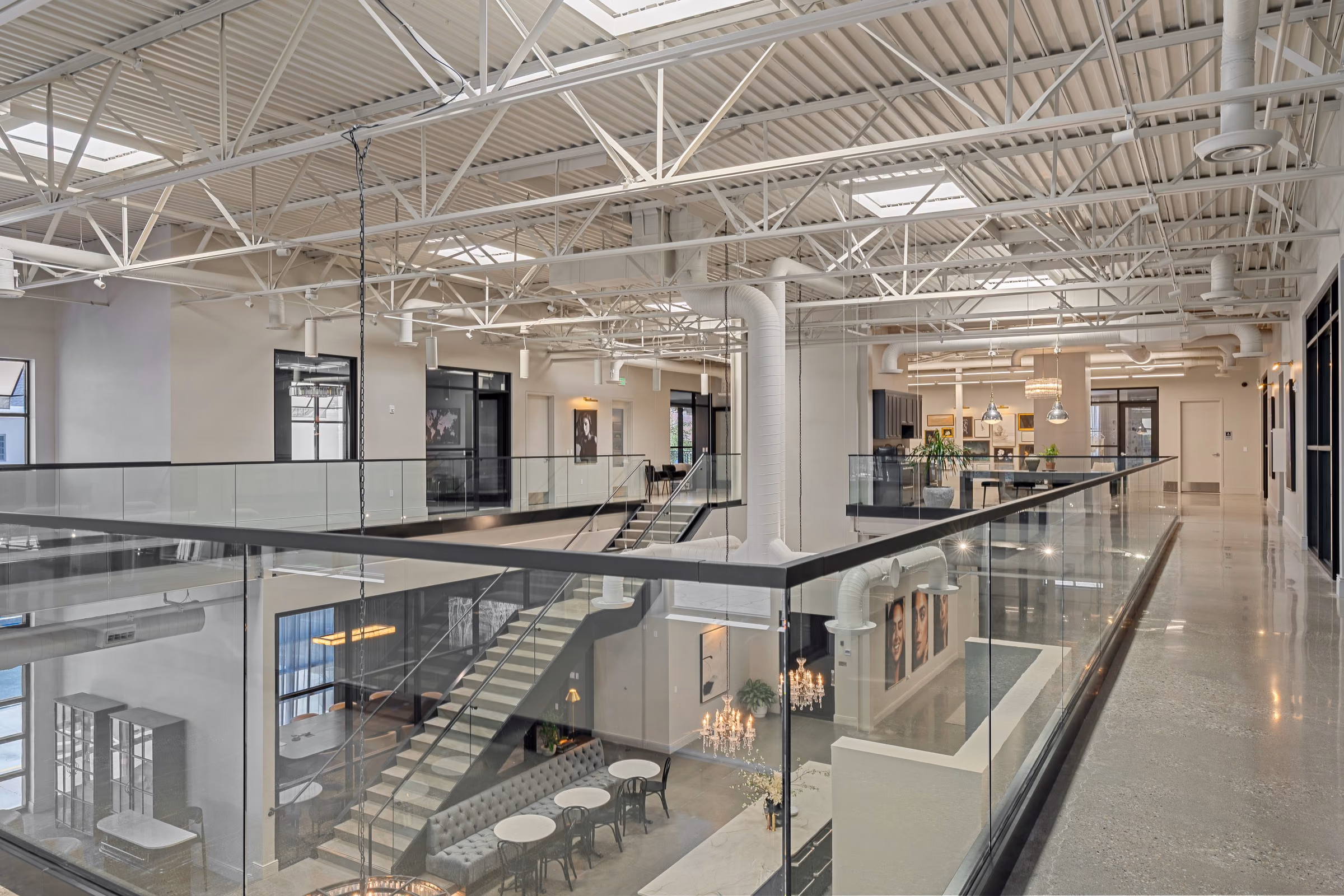View from the mezzanine along a glass guardrail over exposed white steel trusses, round white ductwork, and a stair down to the ground floor, with a polished concrete corridor at right.