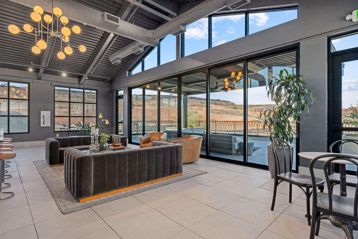 Rooftop lounge under a gabled ceiling with globe chandeliers, channel-tufted grey sofas, tan leather swivel chairs, and floor-to-ceiling sliding glass doors opening to a view of red rock hills.