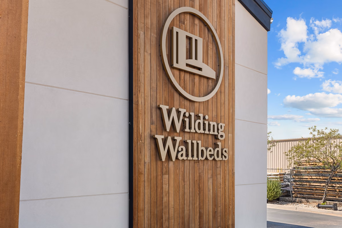 Wilding Wallbeds logo and name in raised metal letters on a cedar-clad sign wall between smooth cream panels, with a blue sky and a stack of lumber behind a fence.