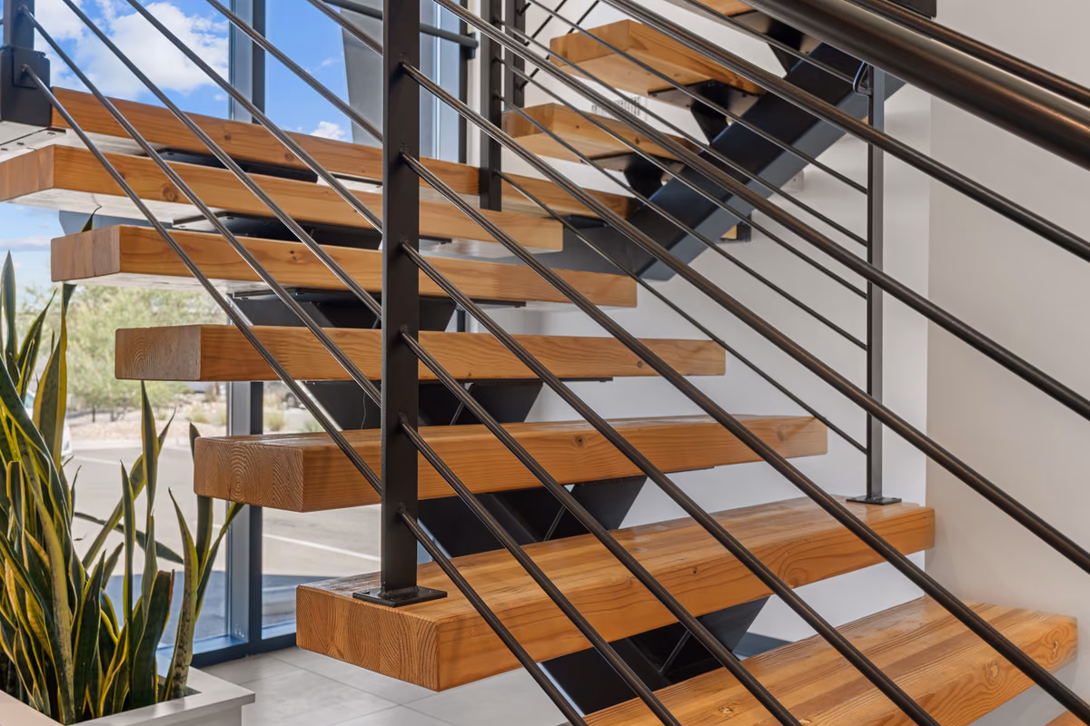 Detail of the timber-tread stair with black horizontal rod railing, a snake plant in a planter, and tall windows behind.